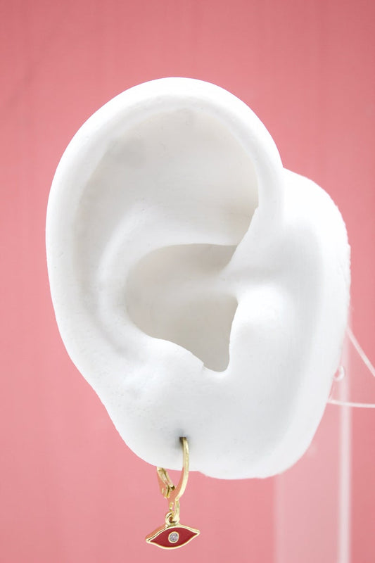 A white mannequin ear displays a gold evil eye charm huggie hoop earring against a pink background.