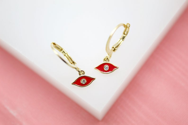 A pair of gold huggie hoop earrings features red enamel evil eye charms with central cubic zirconia stones on a white display surface.