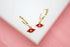 A pair of gold huggie hoop earrings features red enamel evil eye charms with central cubic zirconia stones on a white display surface.