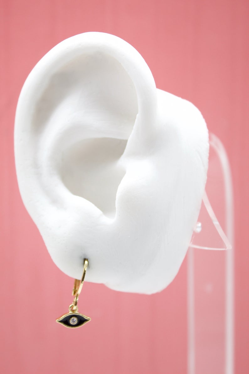 A white display ear showcases a gold evil eye charm hoop earring against a pink background.