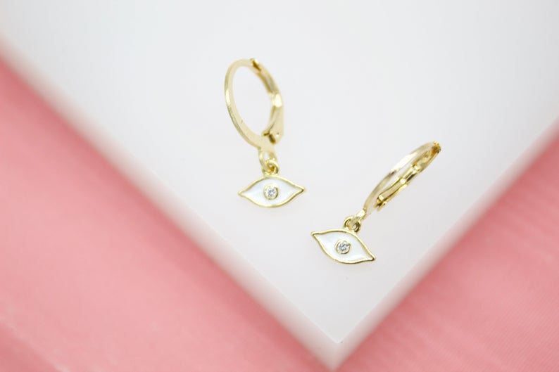 Two gold evil eye hoop earrings with white enamel and sparkling diamond centers rest on a white display block.