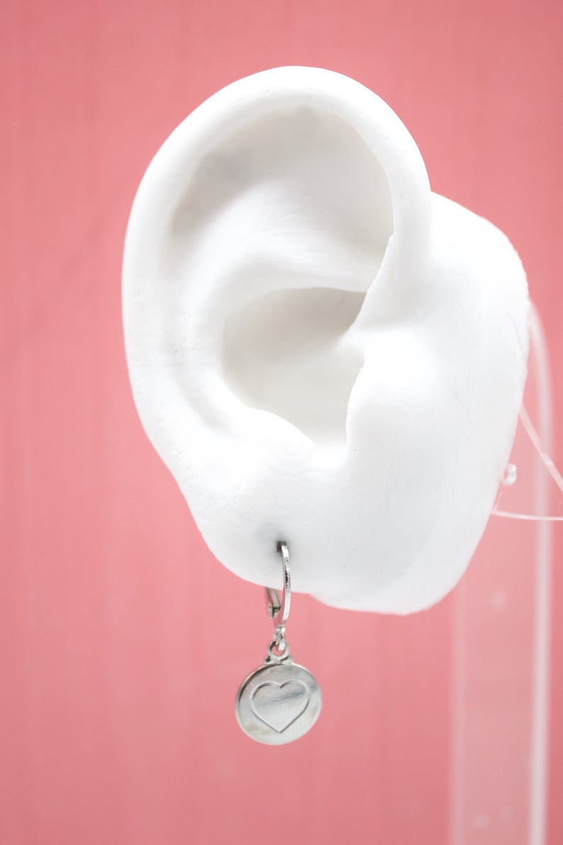 A white mannequin ear displays a silver huggie hoop earring featuring a round pendant engraved with a heart.