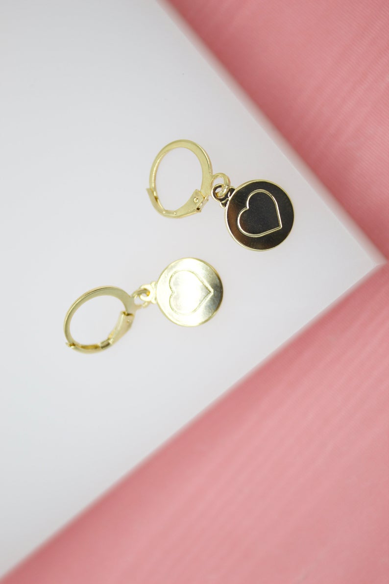 A pair of gold huggie hoop earrings featuring a dangling round pendant with an engraved heart design rests on a white and pink background.