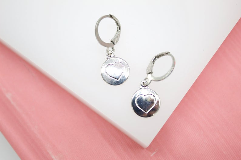 A pair of silver leverback earrings with engraved heart charms rests on a white surface against a pink background.