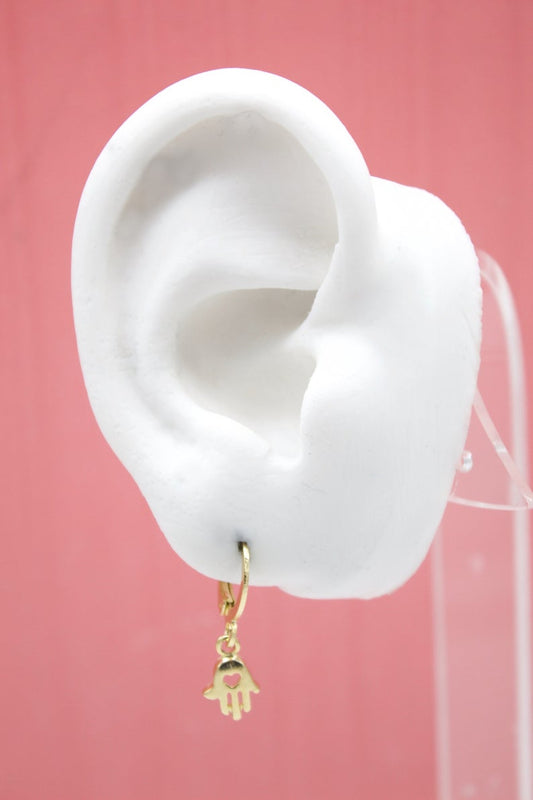 A white mannequin ear displays a gold huggie hoop earring featuring a dangling hamsa hand charm with a tiny heart cutout.