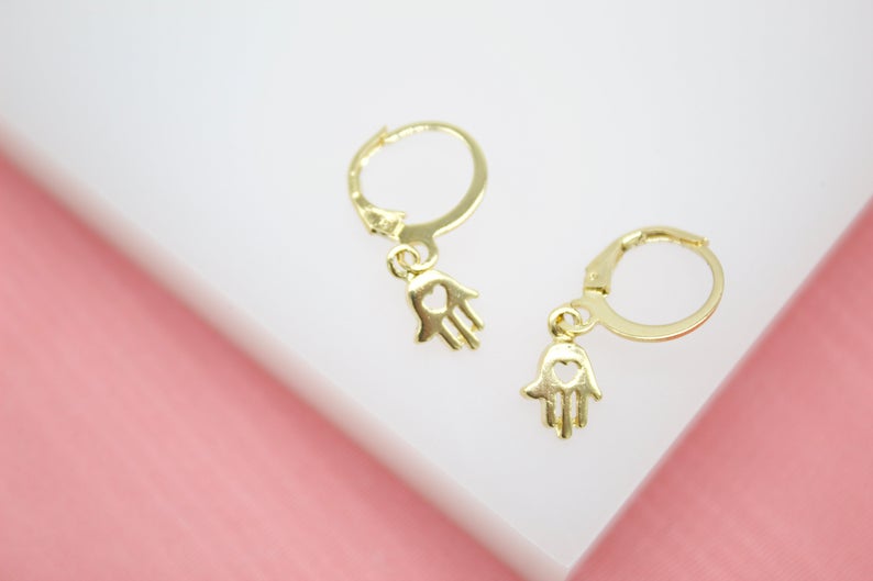 A pair of dainty gold Hamsa hand hoop earrings rests delicately on a soft pink surface.