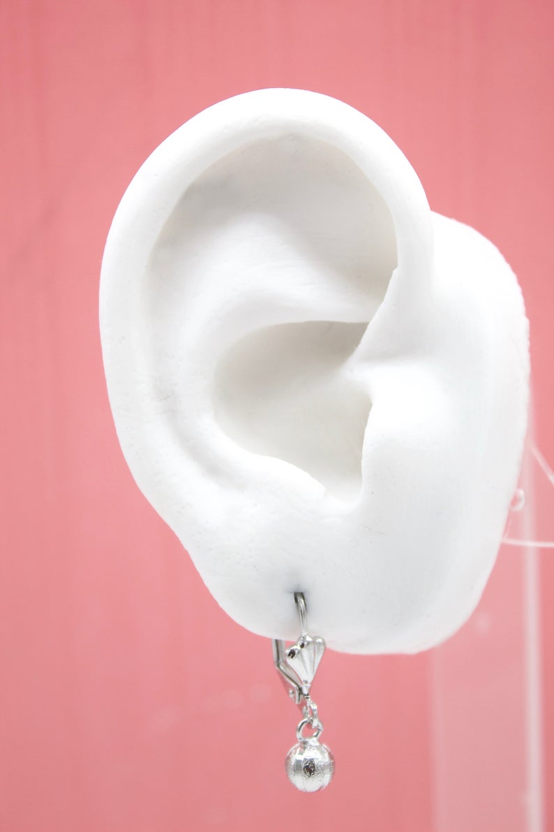 A white mannequin ear displays a silver dangle earring featuring a small heart charm and a round ball pendant against a solid pink background.