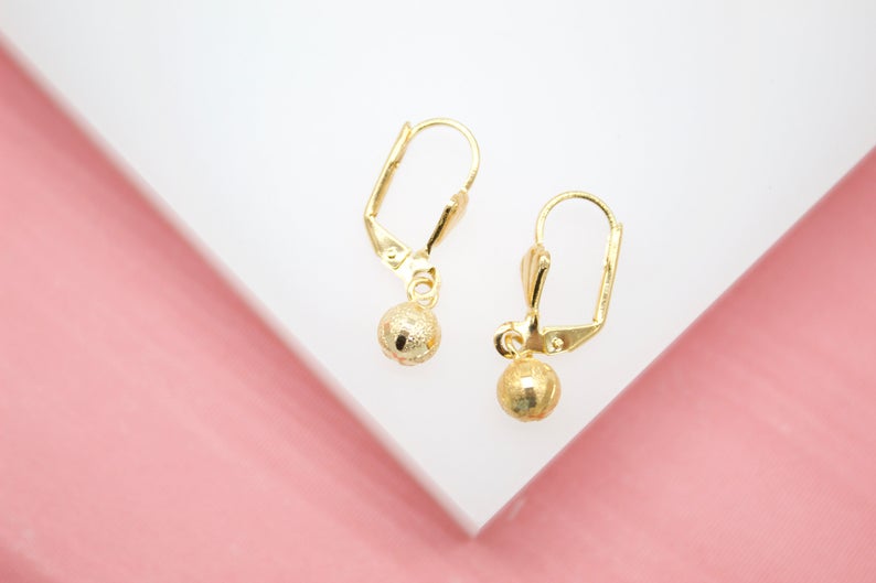 A pair of delicate gold leverback earrings features dangling textured ball charms, displayed on a white surface with a pink background.