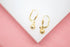 A pair of delicate gold leverback earrings features dangling textured ball charms, displayed on a white surface with a pink background.