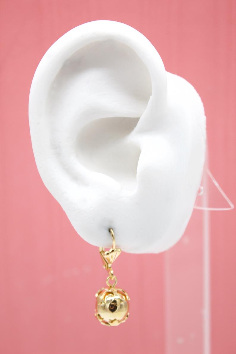 A white ear mannequin displays a dangling gold sphere leverback earring against a solid pink background.