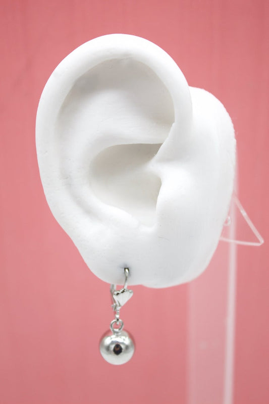 A white mannequin ear displays a silver leverback earring featuring a hanging spherical charm against a solid pink background.