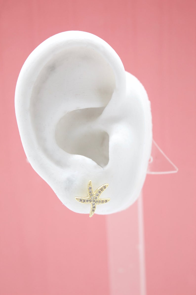 A white display ear model showcases a sparkly gold starfish stud earring against a soft pink background.
