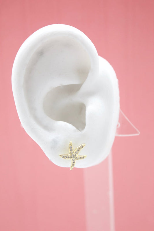 A white display ear model showcases a sparkly gold starfish stud earring against a soft pink background.