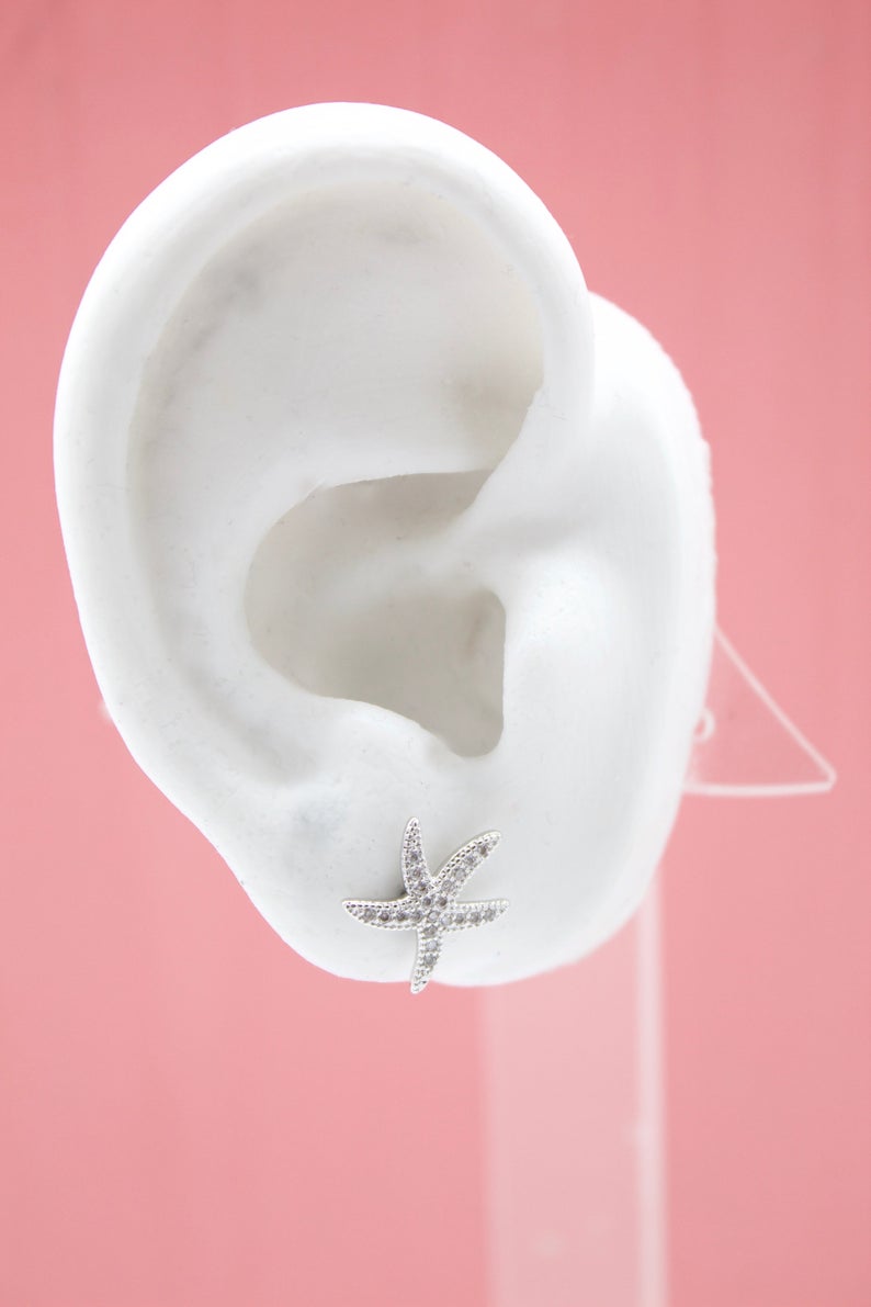 A white mannequin ear displays a sparkling silver starfish stud earring against a pink background.