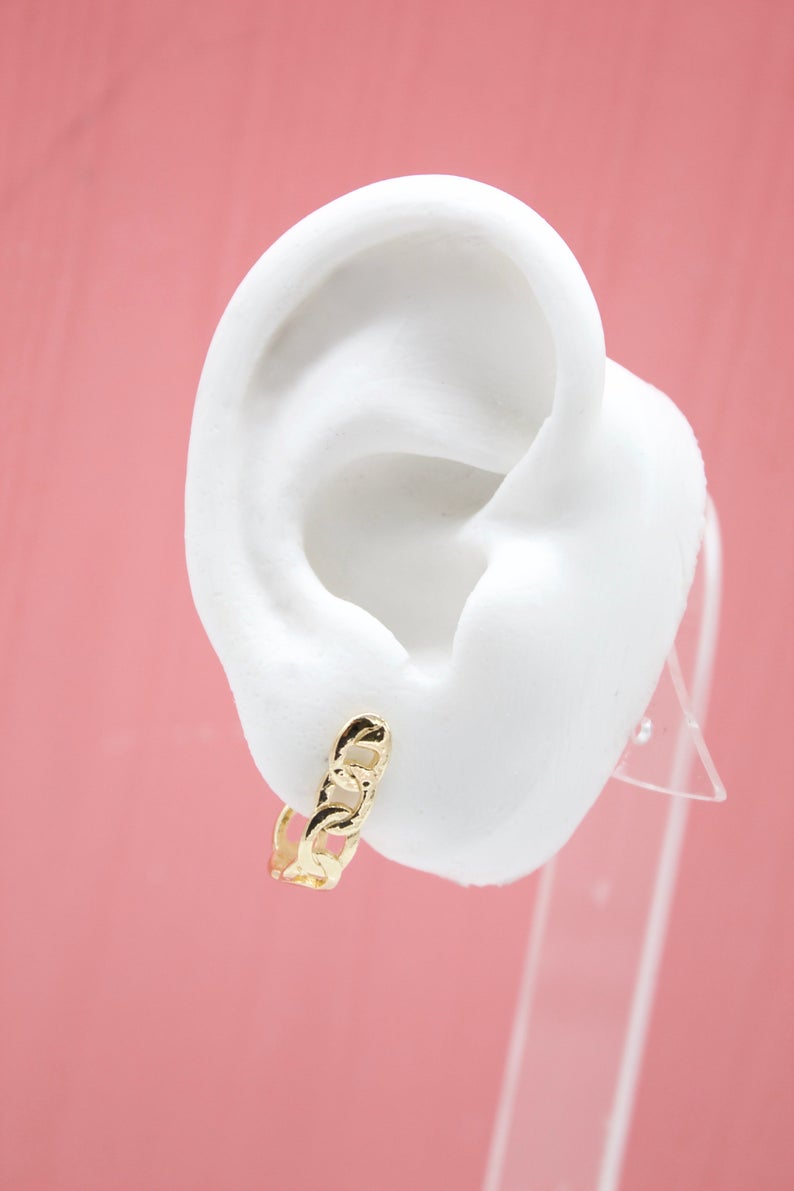 A white mannequin ear displays a gleaming gold chain link huggie hoop earring against a soft pink backdrop.