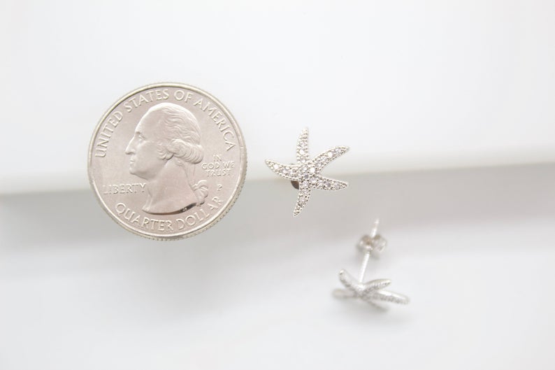 A pair of sparkly pavé-set starfish earrings rests next to a US quarter coin for scale on a white surface.