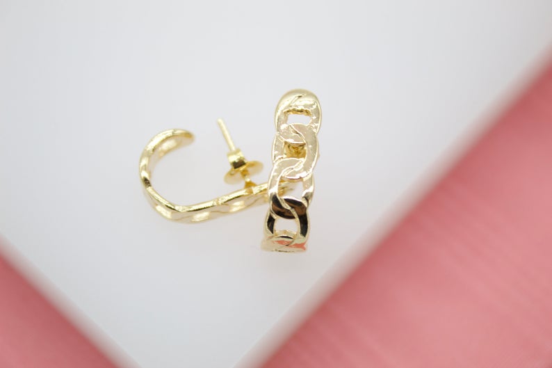A pair of shiny gold chain link stud earrings rests on a clean white surface against a soft pink background.