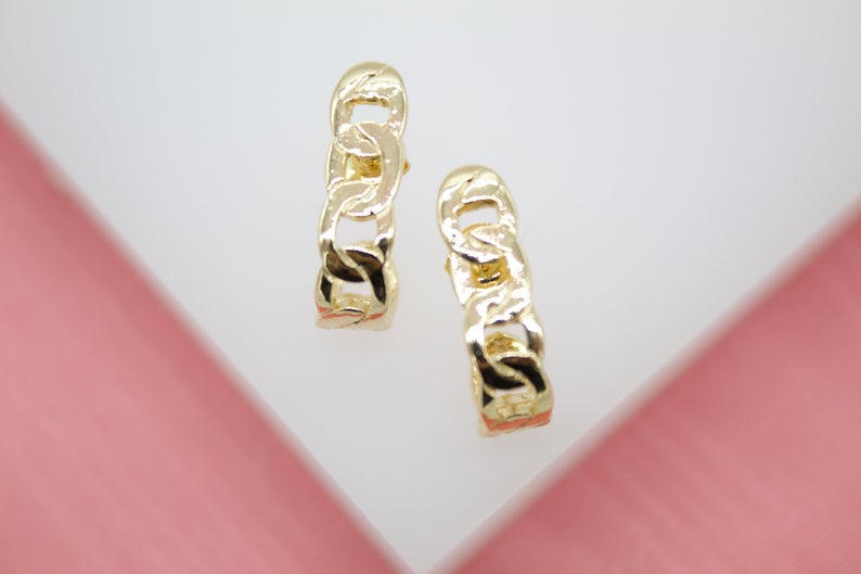 A pair of gold chain-link huggie hoop earrings rests against a white and pink display.