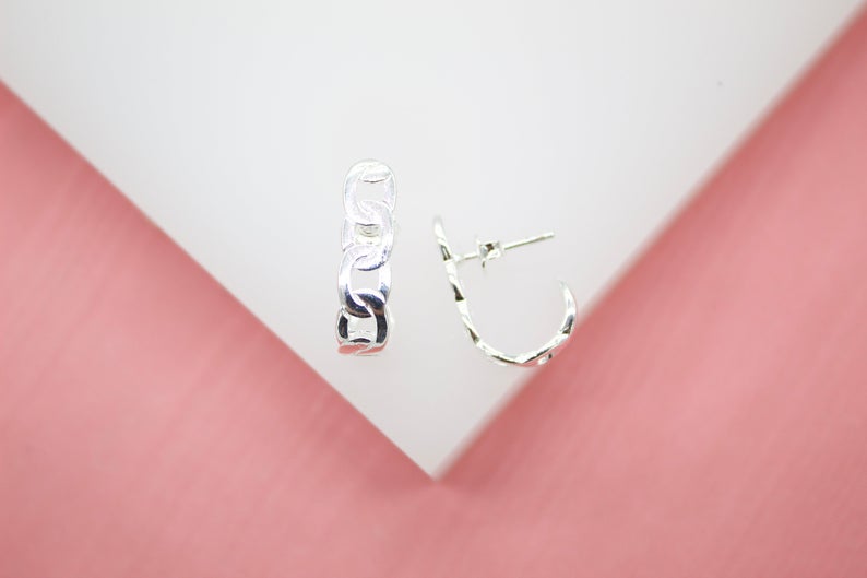 A pair of gleaming sterling silver chain-link hoop earrings rests on a white display surface against a soft pink background.