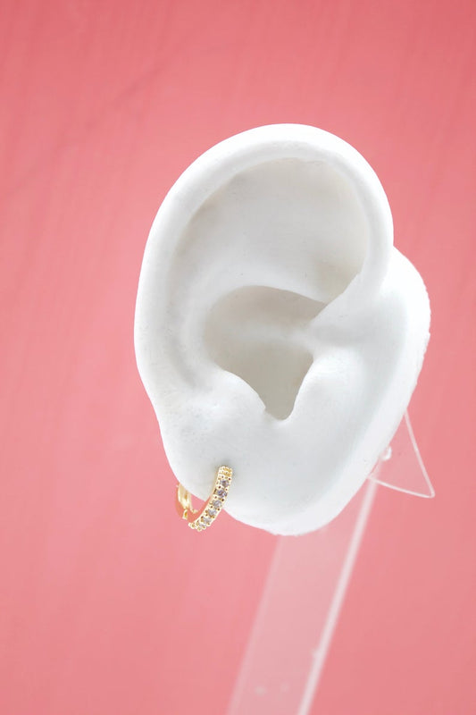 A small gold huggie hoop earring with sparkling pavé crystals adorns the lobe of a white mannequin ear against a solid pink backdrop.