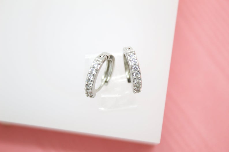 A pair of small silver hoop earrings embellished with sparkling cubic zirconia stones rests on a white display stand.