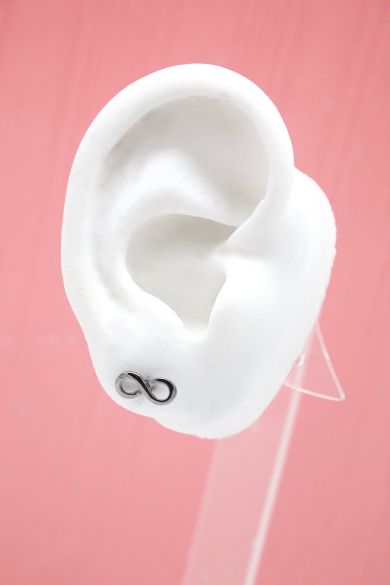 A white mannequin ear displays a delicate silver infinity sign stud earring against a pink background.