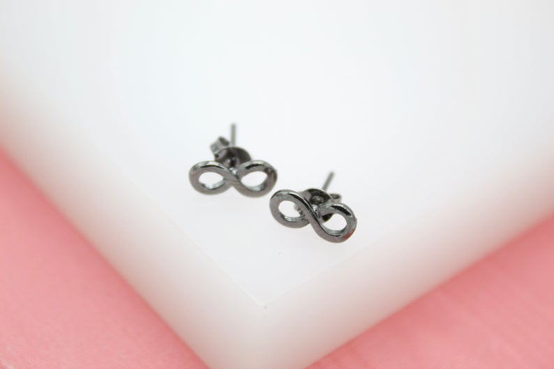 A pair of sleek black infinity symbol stud earrings rests on a clean white display surface against a soft pink background.