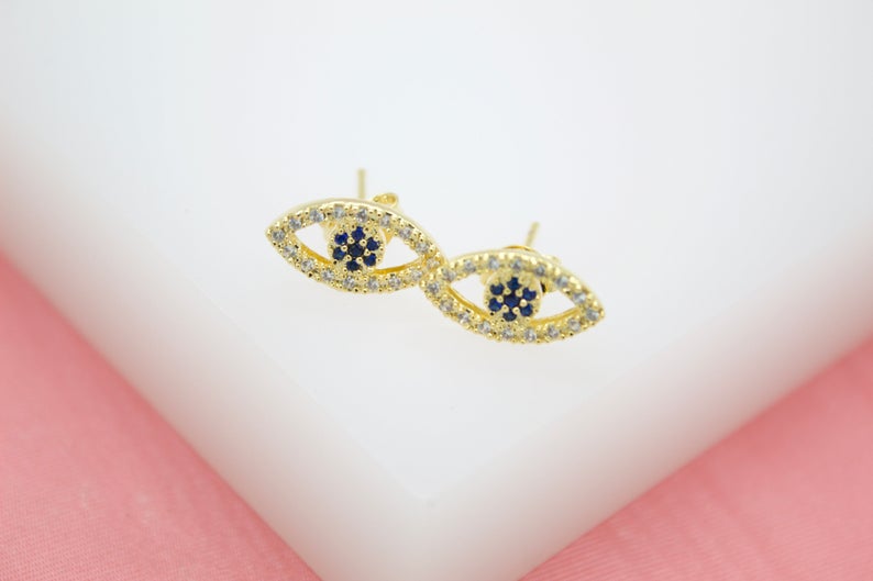 A pair of gold evil eye stud earrings features sparkling pave crystals and dark blue stone centers on a white display surface.