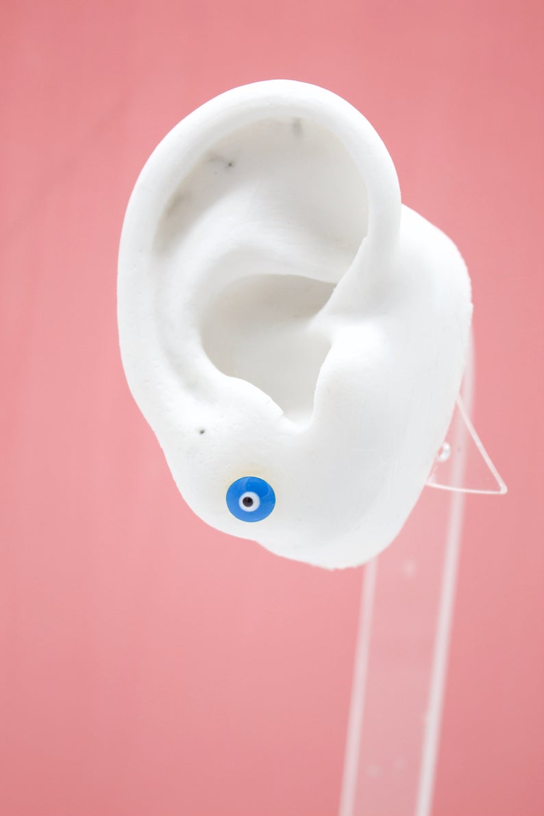 A white mannequin ear on a pink background wears a small blue evil eye stud earring.