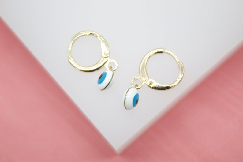 A pair of gold huggie hoop earrings featuring dangling blue and white evil eye charms rests on a white display surface.