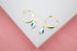 A pair of gold huggie hoop earrings featuring dangling blue and white evil eye charms rests on a white display surface.
