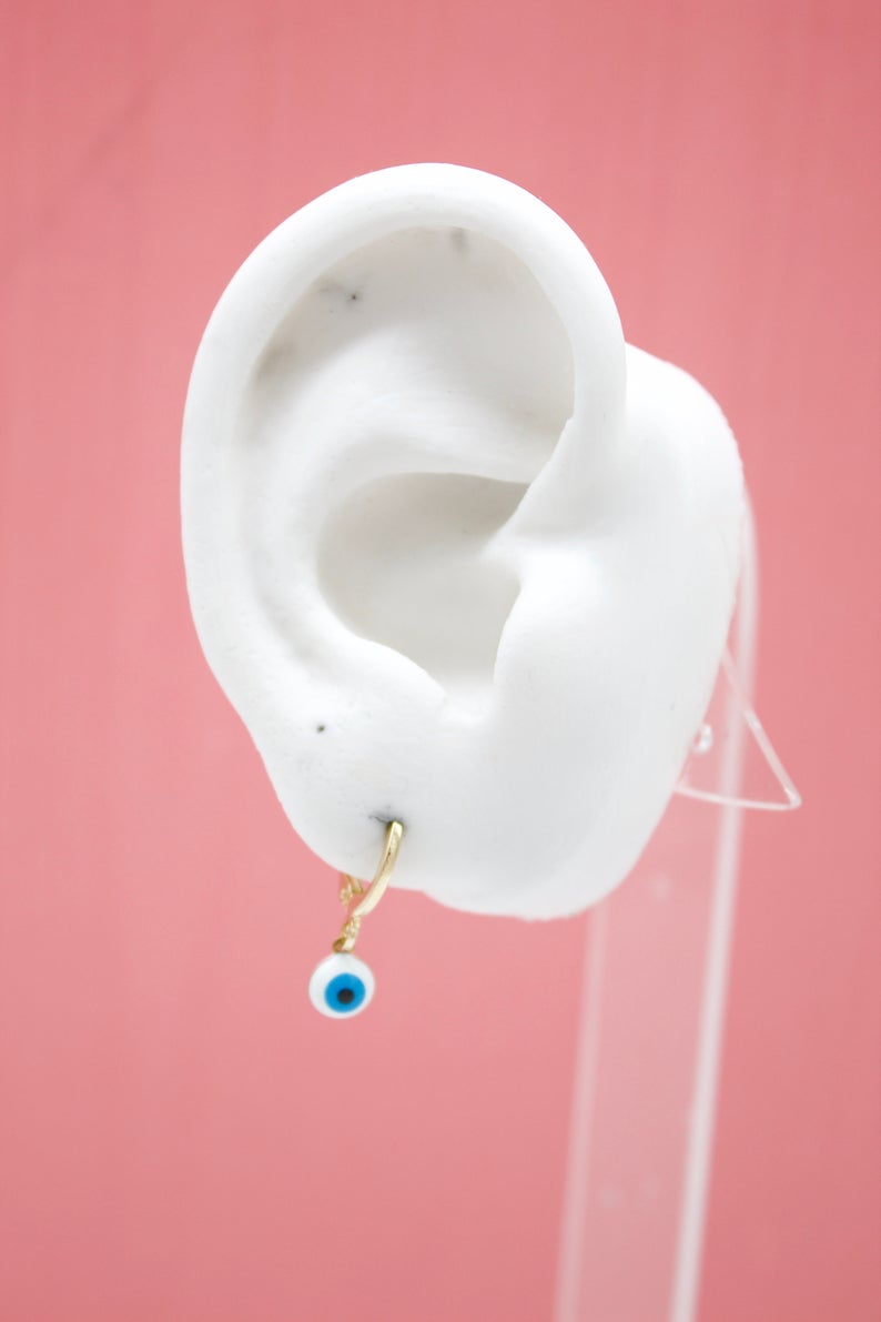 A white mannequin ear on a pink background displays a gold hoop earring featuring a dangling evil eye charm.