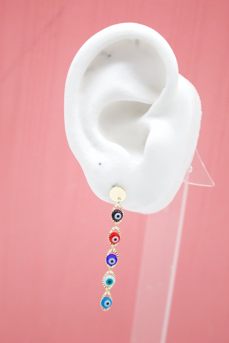 A white mannequin ear displays a dangling gold evil eye charm earring against a pink background.