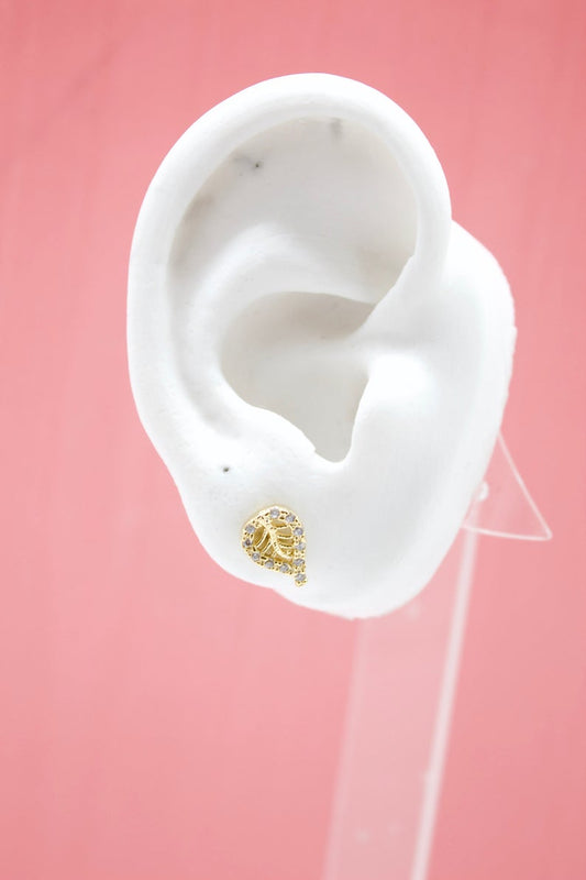 A white mannequin ear displays a sparkly gold leaf stud earring against a pink background.