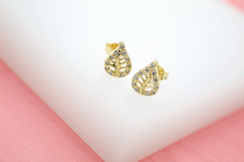 A pair of gold leaf-shaped stud earrings sparkle with embedded cubic zirconia stones on a white and pink display.