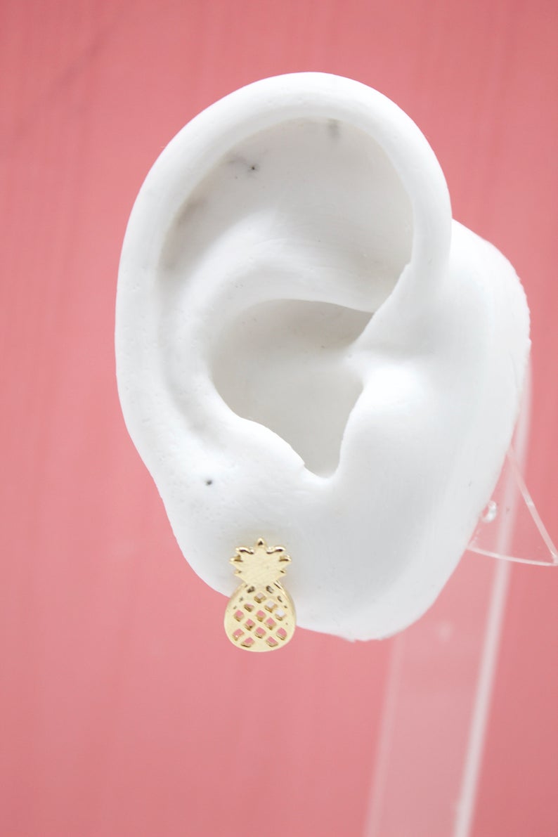 A white display ear showcases a dainty gold pineapple stud earring against a soft pink background.