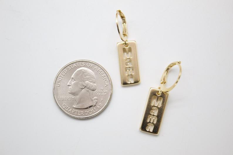 A pair of gold MAMA rectangle dangle earrings lies next to a US quarter coin for scale on a white surface.