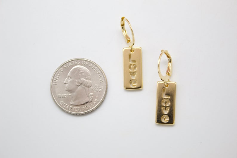 A pair of gold huggie hoop earrings featuring vertical rectangular pendants with the word LOVE engraved vertically, displayed next to a US quarter coin for size comparison on a white background.