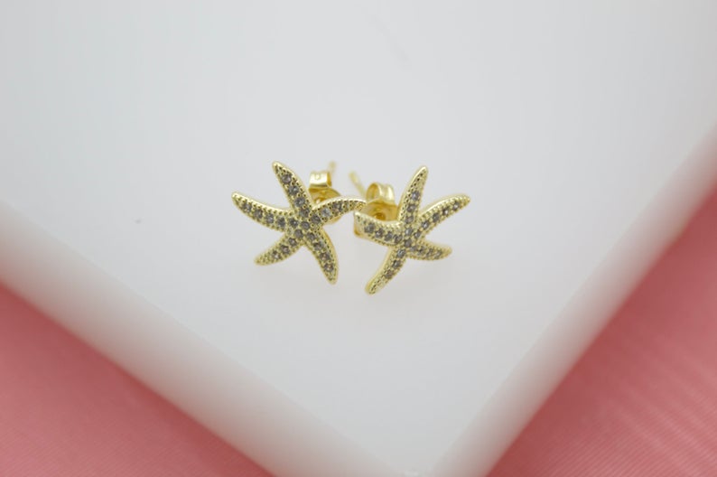 A pair of gold starfish stud earrings encrusted with tiny sparkling rhinestones rests against a white display surface.