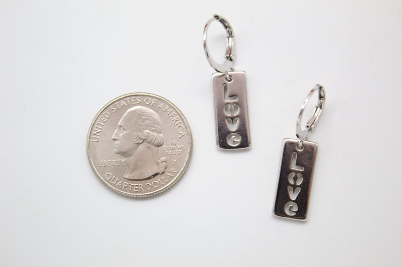 Two silver hoop dangle earrings featuring the word LOVE cutout on rectangular pendants rest next to a US quarter for size comparison on a white surface.