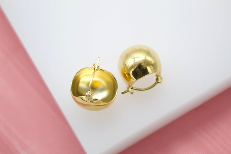 A pair of polished gold dome huggie earrings rests on a white and pink surface.