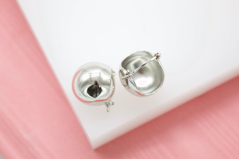 A pair of glossy silver dome hoop earrings rests on a white display against a soft pink background.