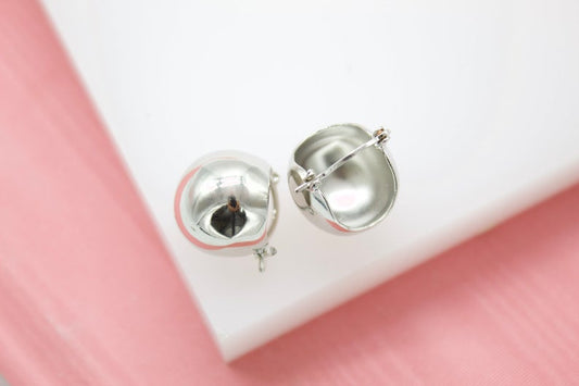 A pair of glossy silver dome hoop earrings rests on a white display against a soft pink background.