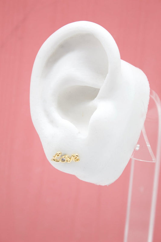A white mannequin ear displays a gold "Love" script stud earring featuring a small sparkling stone.
