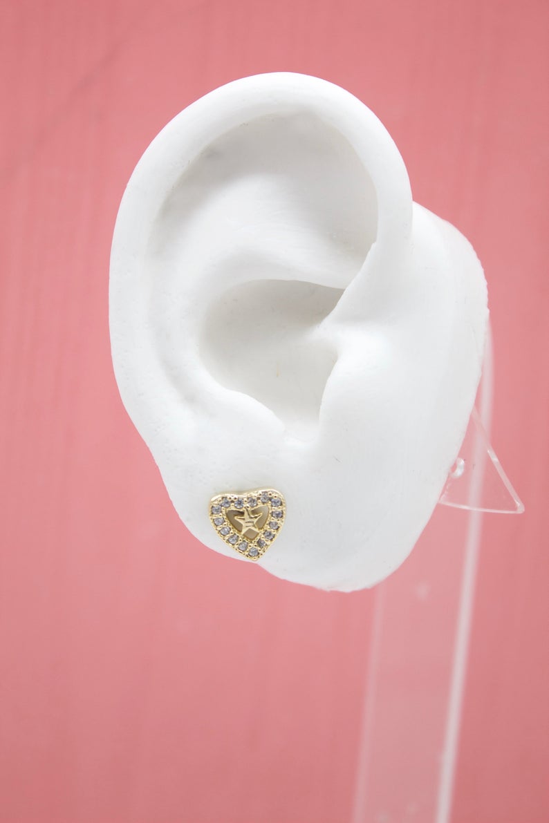 A white mannequin ear displays a gold heart-shaped stud earring encrusted with sparkling cubic zirconia stones and featuring a star design in the center.