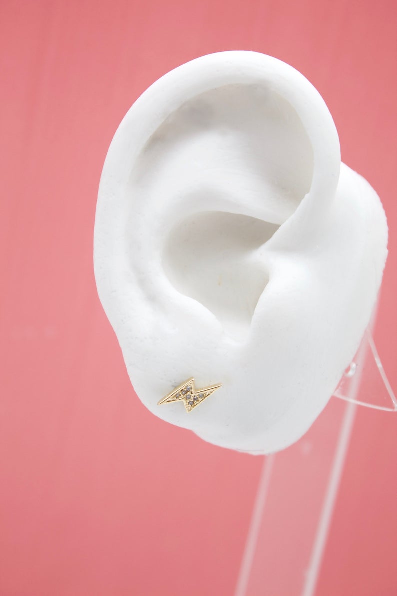 A white mannequin ear displays a sparkling gold lightning bolt stud earring against a pink background.