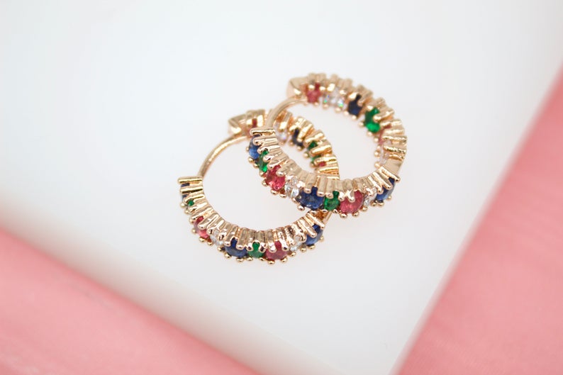 A pair of gold huggie hoop earrings featuring multicolored gemstones rests on a white and pink surface.