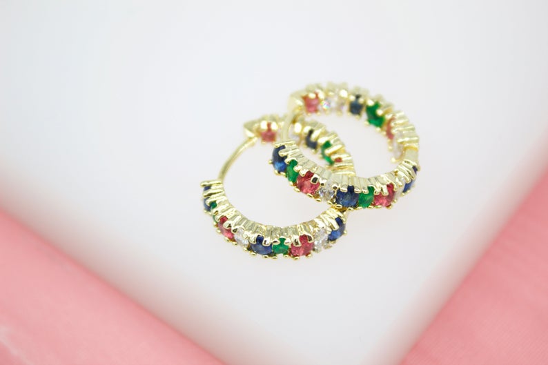 A pair of gold hoop earrings features vibrant multicolored gemstones set along the bands against a soft pink and white background.