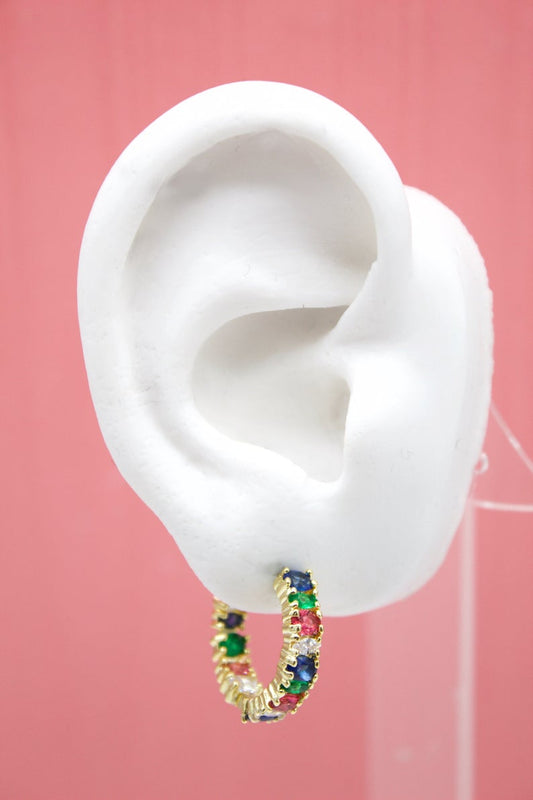A white mannequin ear displays a gold hoop earring studded with colorful multi-gem stones against a soft pink background.