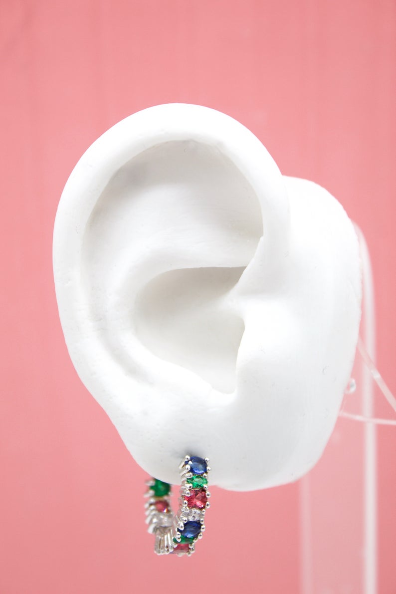 A white ear mannequin displays a colorful multi-stone hoop earring featuring blue, green, and pink gems set in silver against a soft pink background.